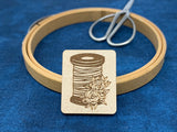 Needle minder featuring a thread spool and flowers. The background shows a wooden embroidery hoop and a pair of silver scissors.