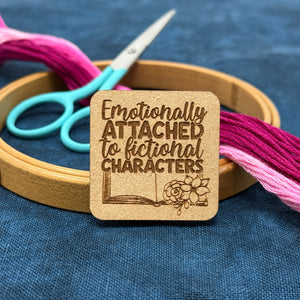 Photo of a needle minder with the quote, "Emotionally attached to fictional characters" with an open book and flowers.