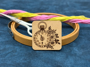 Photo of a needle minder featuring a pocket watch surrounded with flowers.