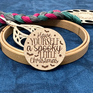 Round laser cut wood needle minder saying "Have Yourself A Spooky Little Christmas. Wood embroidery hoop, gold scissors, thread keep embroidery floss organizer, and embroidery floss are in the background.