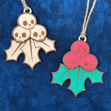 Hand-painted and unpainted Creepy Christmas Skull Holly Berry laser cut wood ornament laying on a blue background.